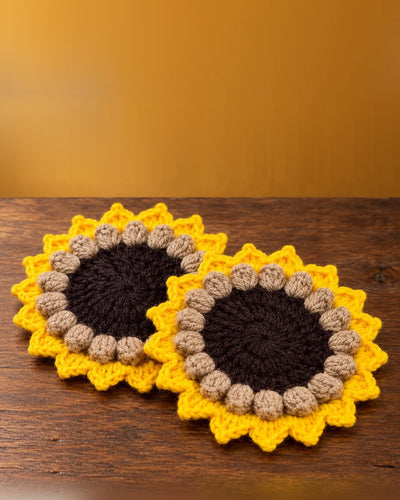 Two crochet sunflower coasters with brown centres, taupe bobble rings and yellow petals on a wooden table